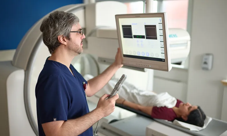 A medical professional operates a touchscreen control panel while a patient lies on a scanning bed inside a medical imaging machine in a clinical setting.