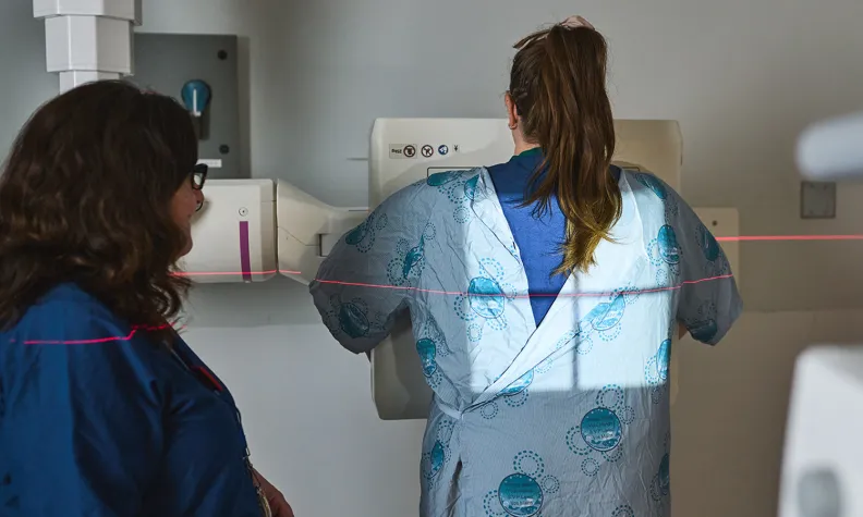 Une personne vêtue d’une jaquette d’hôpital se tient debout face à un appareil d’imagerie médicale. Une technicienne est à ses côtés. Une ligne laser rouge visible dans son dos indique que la patiente subit un examen diagnostique.