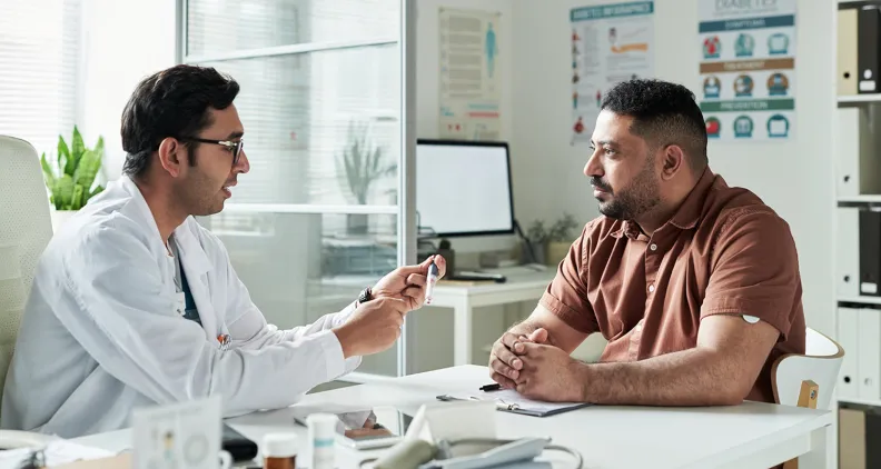 Un médecin en blouse blanche est assis à un bureau et explique quelque chose à un patient vêtu d’une chemise marron. Du matériel médical, des documents et des affiches sur la santé sont visibles en arrière-plan.