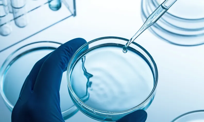 Une main gantée tient une boîte de Pétri contenant un liquide transparent. Une pipette est au-dessus de la boîte de Pétri. Elle libère une goutte. Des tubes de verre et des reflets sont visibles à l’arrière-plan.