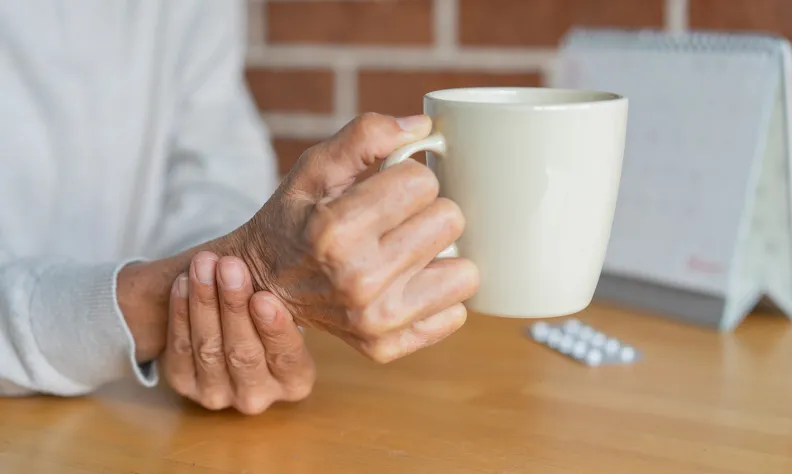Une personne âgée tient une tasse blanche d’une main tout en soutenant son poignet de l’autre; un calendrier et des comprimés sont visibles sur une table en bois à l’arrière-plan.