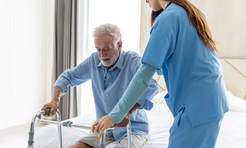 An elderly patient uses a walker to stand up from a bed, assisted by a healthcare worker in blue scrubs in a bright, well-lit room.