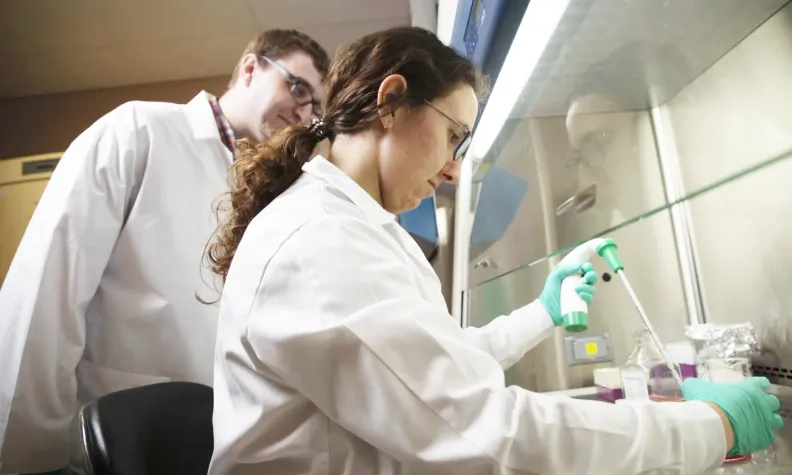 Two individuals in lab coats work at a laboratory station, with one using a pipette inside a fume hood.