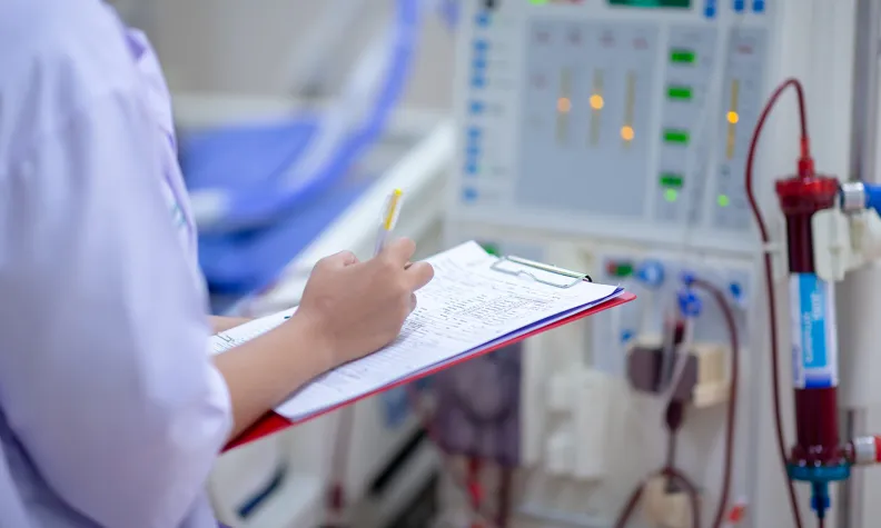 A healthcare worker in a lab coat writes notes on a clipboard while standing next to medical equipment with illuminated digital displays and tubing.