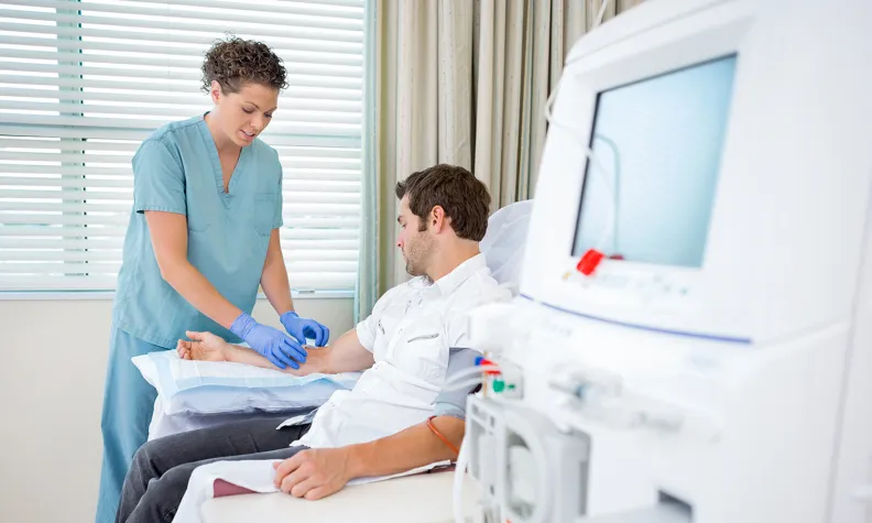 A nurse wearing scrubs and gloves prepares a patient for a medical procedure in a clinic room, with dialysis equipment visible in the foreground.