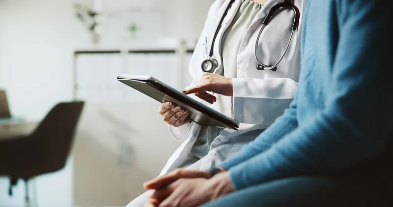 A healthcare professional wearing a stethoscope reviews information on a digital tablet while sitting beside a patient in a medical office.