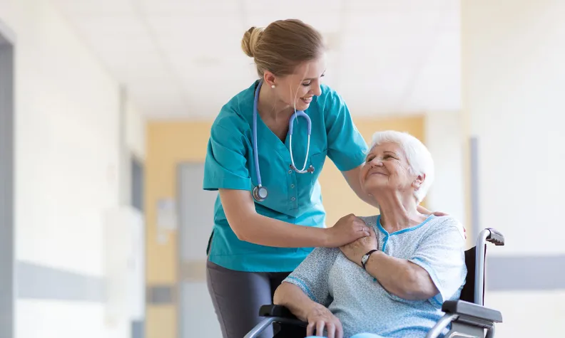 A healthcare worker in blue scrubs smiles and gently talks to an elderly patient sitting in a wheelchair in a bright hallway, showing care and support.