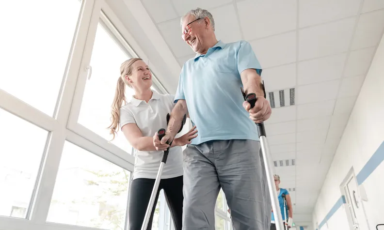 Un patient âgé souriant utilise des béquilles pour marcher dans un couloir. Il est accompagné d’une travailleuse de la santé qui lui offre du soutien dans le cadre d’une séance de physiothérapie ou de réadaptation.
