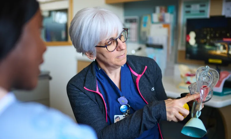 A healthcare professional with gray hair and glasses explains a medical diagram of the female reproductive system to a patient in a clinical setting.