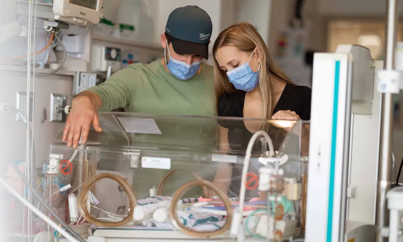 Two people wearing masks look down at a newborn baby inside an incubator in a hospital neonatal intensive care unit, surrounded by medical equipment.
