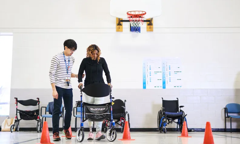 Une personne marche à l’aide d’une marchette en suivant un parcours de cônes orange dans un gymnase. La personne est soutenue par une autre personne à ses côtés. Des fauteuils roulants sont alignés le long du mur sous un panier de basketball.