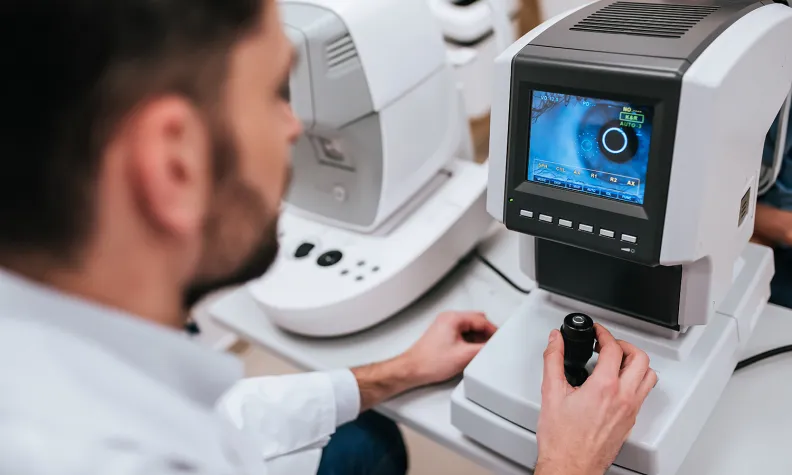 Salle d’examen en ophtalmologie où un clinicien utilise un appareil d’imagerie rétinienne, observant une image détaillée de l’œil sur l’écran de l’appareil pendant qu’un patient est positionné à l’instrument pour un examen de la vue.