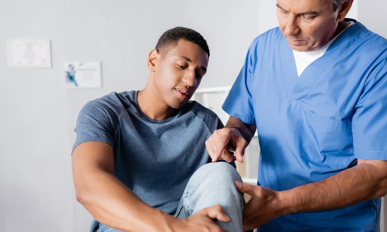 A healthcare professional in blue scrubs examines a patient's knee, gently supporting the leg while assessing the joint.