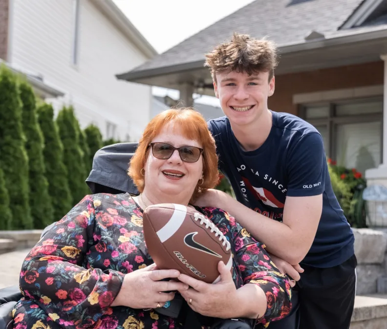 A woman sitting in a wheelchair holds a football while a young man stands close beside her with his arm around her shoulders. They are outside in a residential neighborhood, in front of a house with greenery in the background.