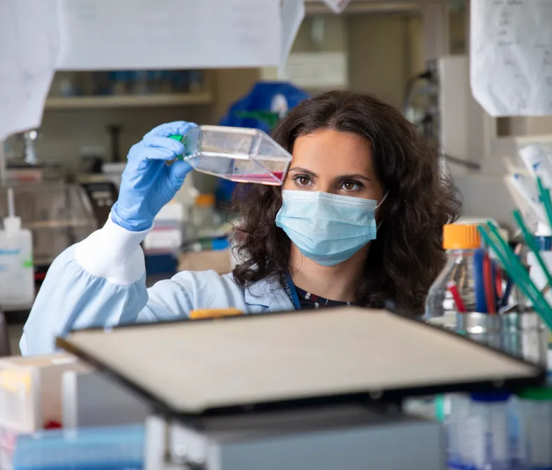 In a laboratory setting, a researcher wearing a lab coat and blue gloves holds up a container of pink liquid media and examines it closely. Scientific equipment, bottles, and tools are visible throughout the workspace.
