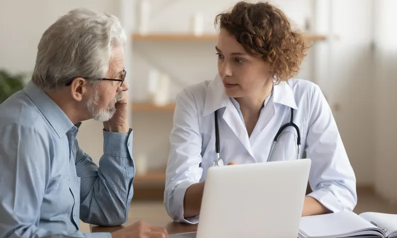 A healthcare professional speaks with an older patient at a desk, reviewing information together on a laptop and in an open notebook.
