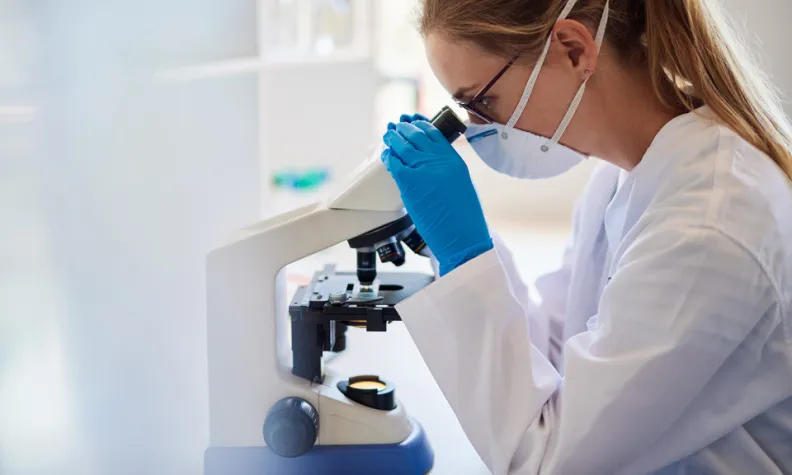 Un professionnel de laboratoire portant une blouse blanche et des gants bleus observe un microscope sur un poste de travail dans un laboratoire clinique ou de recherche. 