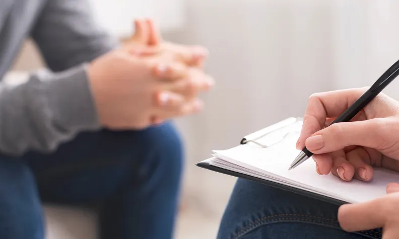 A person sits with hands clasped while another holds a clipboard and writes with a pen, suggesting a counseling or therapy session.