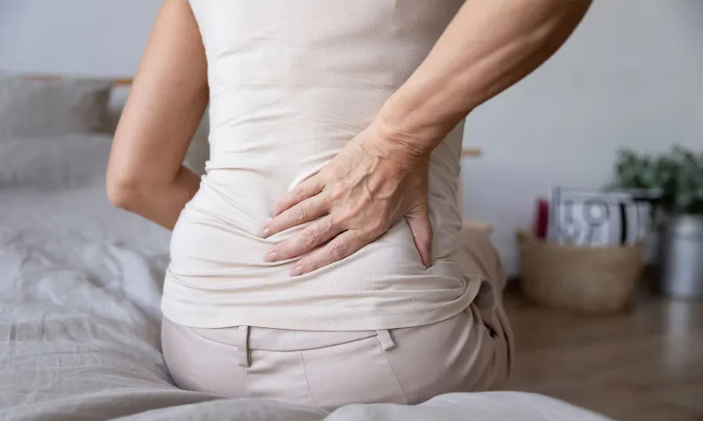 Une personne vêtue de vêtements pâles est assise sur le bord d’un lit. Elle se tient le bas du dos d’une main et semble souffrir de douleurs dorsales.