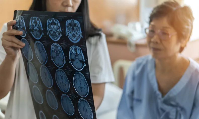 A healthcare professional holds up and examines brain scan images while an older person in a hospital gown sits next to them, watching attentively in a medical office setting.