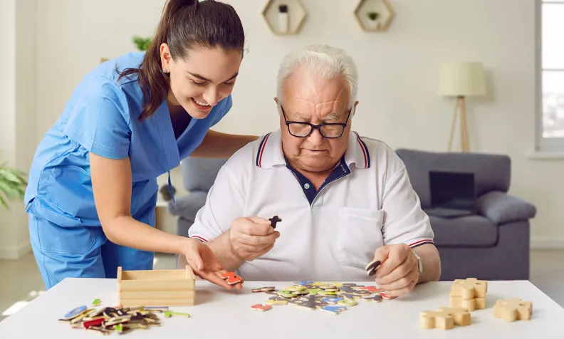 A caregiver in blue scrubs smiles and assists an elderly person with glasses as they work on a puzzle at a table in a bright, cozy living room.