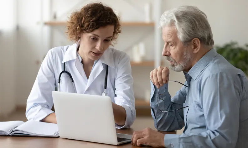 A healthcare professional in a white coat shows information on a laptop to an older person with gray hair and a beard. They sit at a desk, appearing focused and engaged in discussion.
