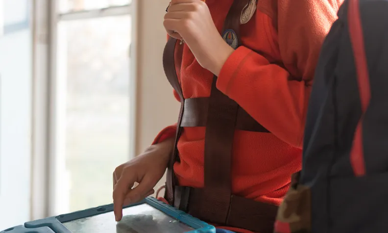 A person in an orange fleece jacket uses a tablet with one hand while chewing on a pencil. The person is wearing a harness to carry the tablet and standing near a window with daylight streaming in.