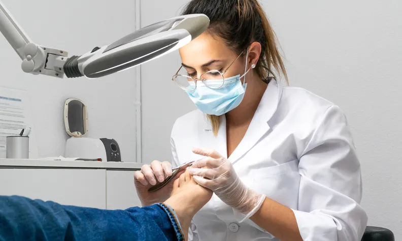 A podiatrist wearing a mask, gloves, and white coat uses a tool to treat a patient’s foot under a bright examination lamp in a medical office.