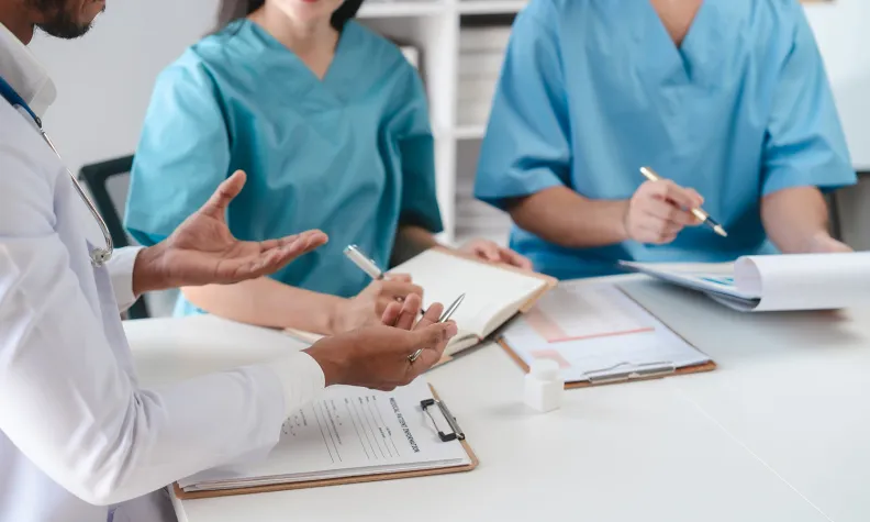Three healthcare professionals in scrubs sit around a table, discussing notes and holding pens, with medical documents and clipboards in front of them.