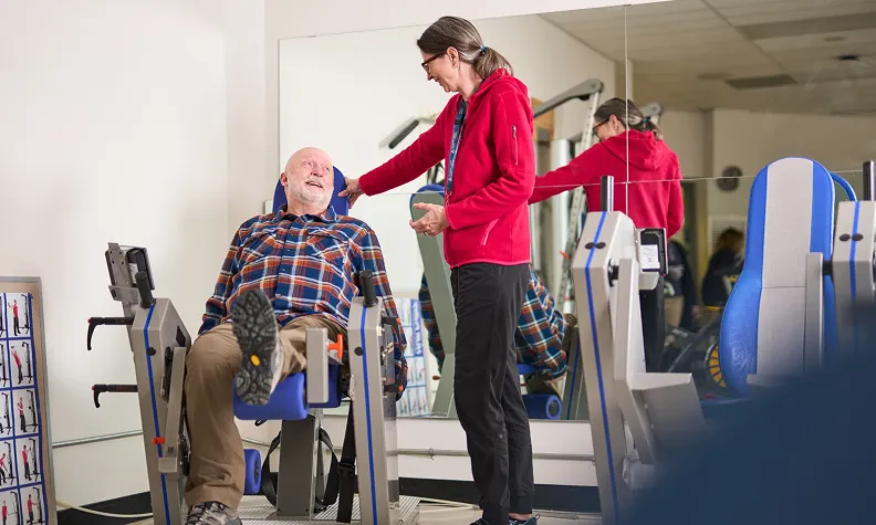 An older person uses a leg exercise machine while a person in a red hoodie smiles and in a gym or physical therapy room.