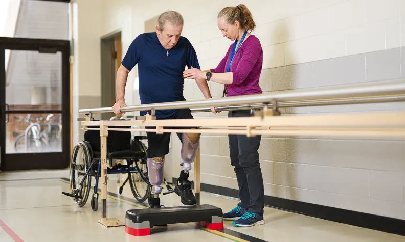 A physical therapist assists an older person with prosthetic legs as they practice walking using parallel bars in a rehabilitation facility. A wheelchair is positioned nearby.