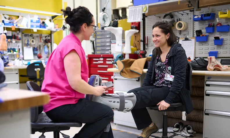 Two people sit facing each other in a workshop. One is wearing a prosthetic leg and smiling, while the other, in a pink shirt, inspects or adjusts the prosthetic. Tools and prosthetic parts are visible in the background.