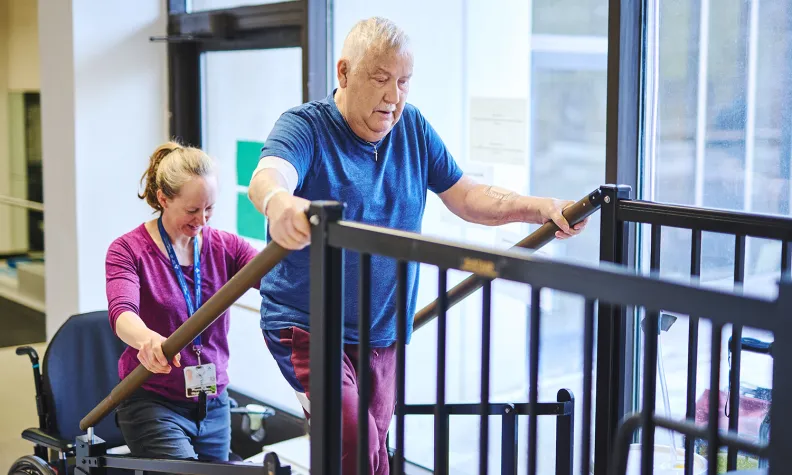 A physical therapist assists an older adult as he practices climbing a small set of therapy stairs, using handrails for support in a rehabilitation facility 