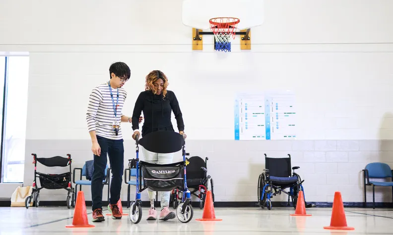 A person uses a walker to navigate an obstacle course marked by orange cones in a gym, assisted by a person beside them. Several wheelchairs are positioned along the wall under a basketball hoop.