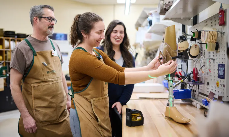 Three people in aprons stand at a workbench in a workshop, smiling and working on assembling a prosthetic leg. Tools and materials are organized on the wall and shelf in the background.