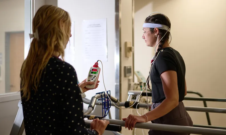 A person walks on a treadmill while wearing sensors and a headband, monitored by another person holding a device with a display, inside a medical or research facility.