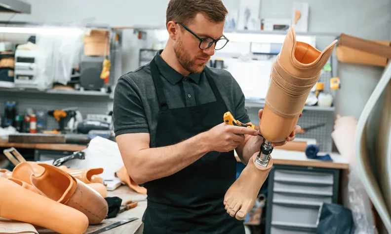 A person wearing glasses and an apron works on assembling a prosthetic leg in a workshop filled with tools and prosthetic limbs.