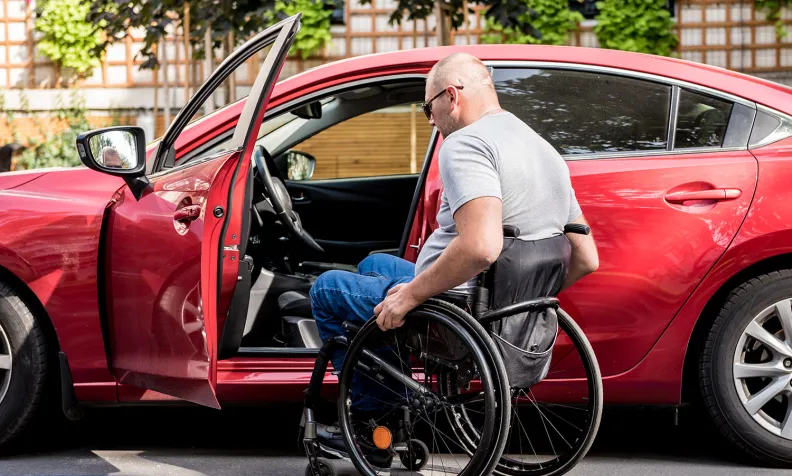 A person in a wheelchair transfers themself into the driver’s seat of a red car parked in an accessible parking spot, with the driver’s door open and greenery visible in the background.