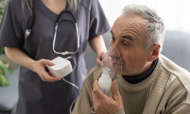 An older person wearing a beige sweater uses a nebulizer mask while sitting. A healthcare professional stands beside them holding the nebulizer machine.