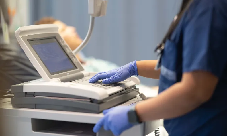 A healthcare professional wearing blue scrubs and gloves operates an ultrasound machine, with a patient lying on an examination table in the background.