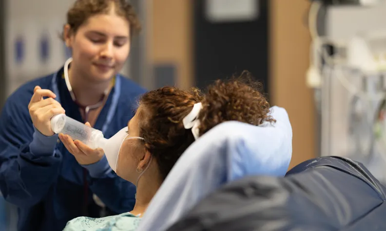 A healthcare worker assists a patient sitting in a hospital chair by holding an oxygen mask to their face. The scene takes place in a medical setting with blurred background equipment.