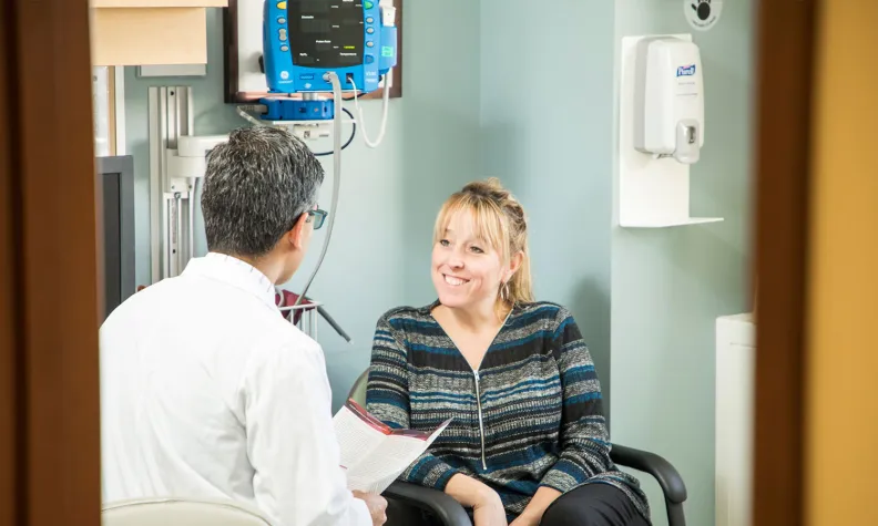 Dans un environnement médical, un professionnel de la santé tenant un document est assis en face d’une personne également assise et discute avec celle-ci.