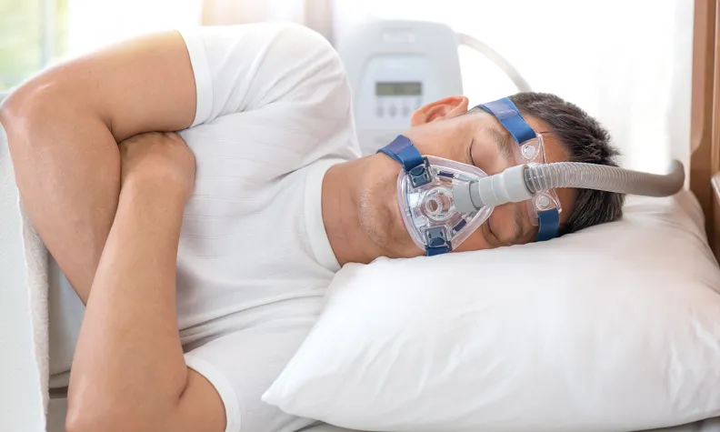 A person sleeps on their side while wearing a CPAP mask connected to a sleep apnea machine.