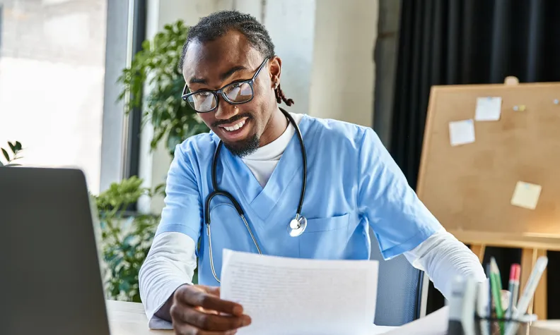 A healthcare professional wearing scrubs and a stethoscope reviews a document while sitting at a desk with a laptop in a clinical or office setting.