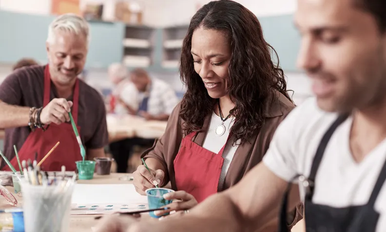 Des adultes portant des tabliers sont assis à une table dans une salle de cours d’art, souriant et peignant avec des pinceaux et des pots de peinture.