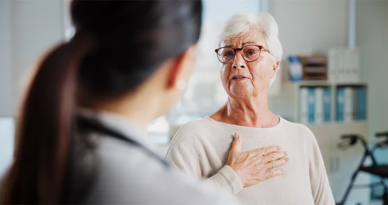An older patient wearing glasses touches their chest while speaking to a healthcare professional in a medical office. 