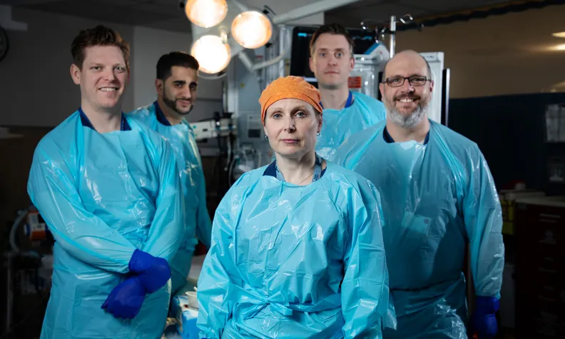 Cinq professionnels de la santé portant des tenues bleues, des gants bleus et des blouses de protection bleues sont debout ensemble dans un environnement hospitalier.