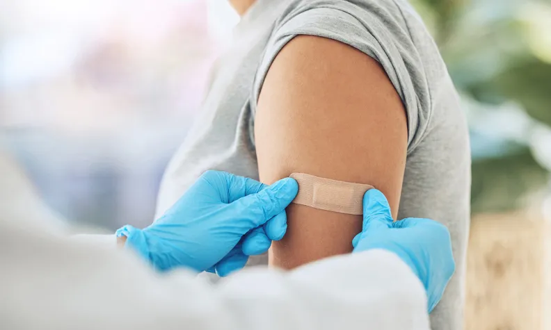 A healthcare professional wearing blue gloves places a bandage on a patient’s upper arm after an injection or vaccination.