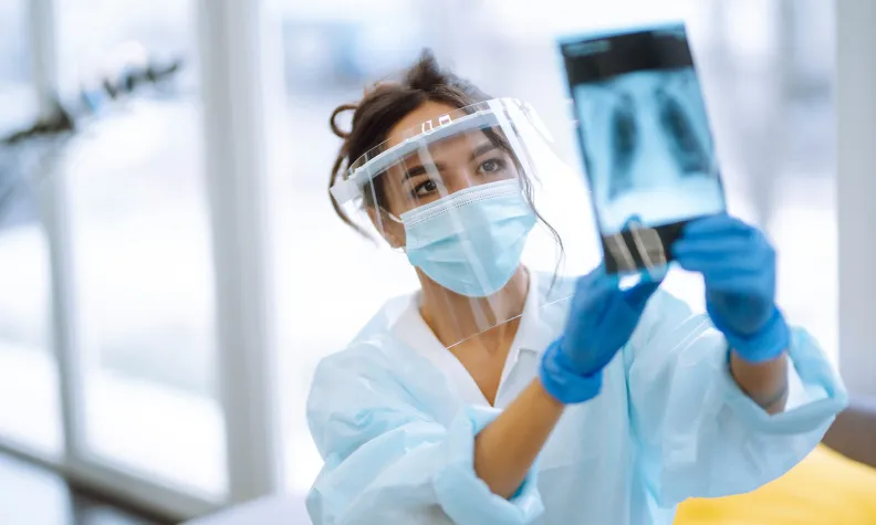 A healthcare worker wearing a mask, face shield, gloves, and gown examines a chest X-ray while standing in a bright medical facility.