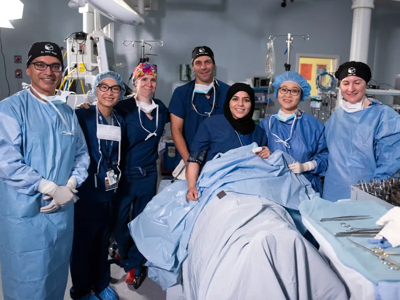 A group of six medical professionals stands around another health-care worker acting as a patient on an operating table, all wearing scrubs, masks around their necks, and caps.